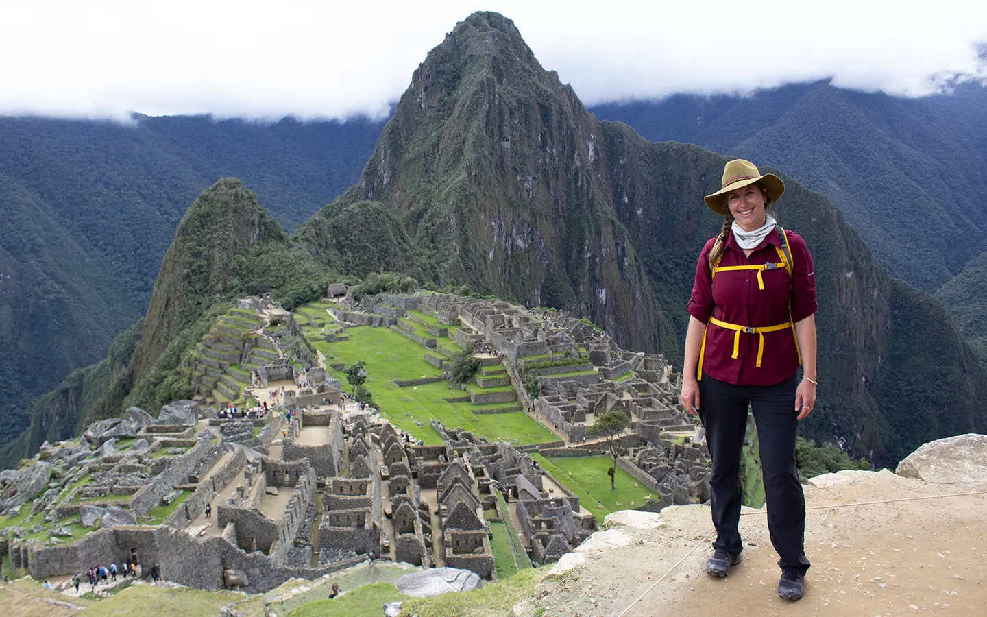 A woman in a red shirt, yellow backpack straps and a safari style hat stands on a ledge above Machu Picchu