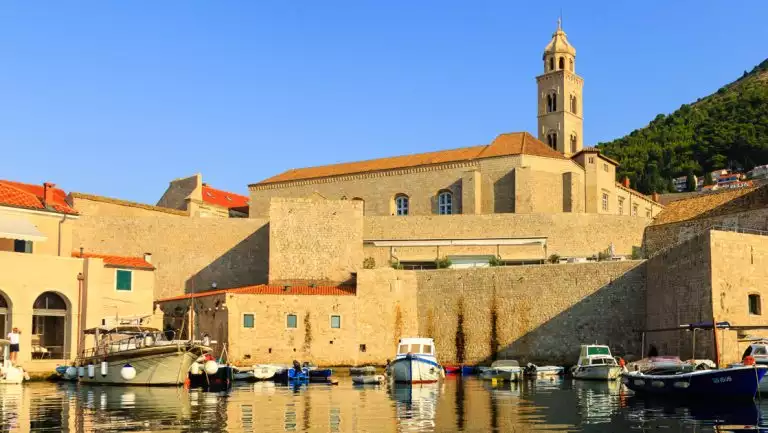 Small fishing boats of various colors sit in glassy harbor water by tan stone buildings, seen on an Ancient Med Odyssey cruise.