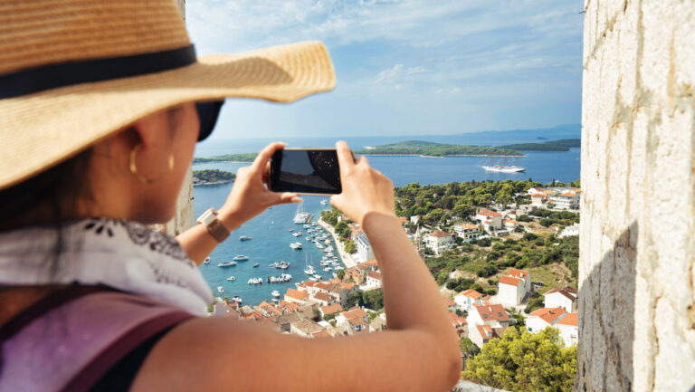 Woman in woven sunhat holds phone to photograph Hvar, Croatia with beige stone buildings, terracotta roofs & green trees.