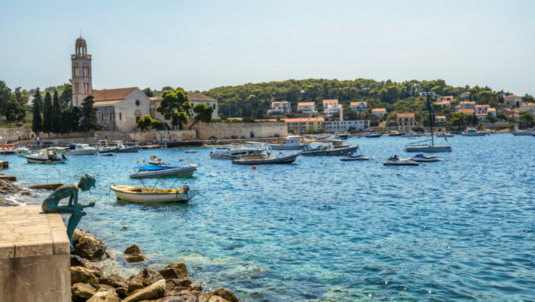 Weathered stone statue of a person sits overlooking teal sea & small fishing boats on moorings beside sunny, seaside town.