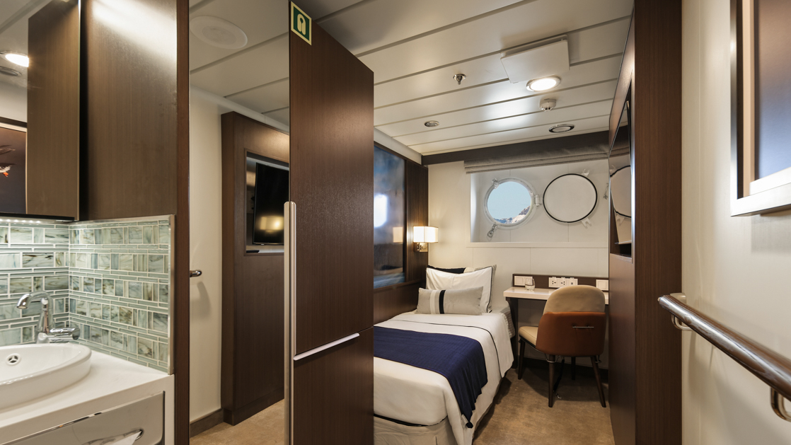 Single Stateroom on Douglas Mawson ship with twin bed in white linens, dark wood walls, porthole & wildlife photos.