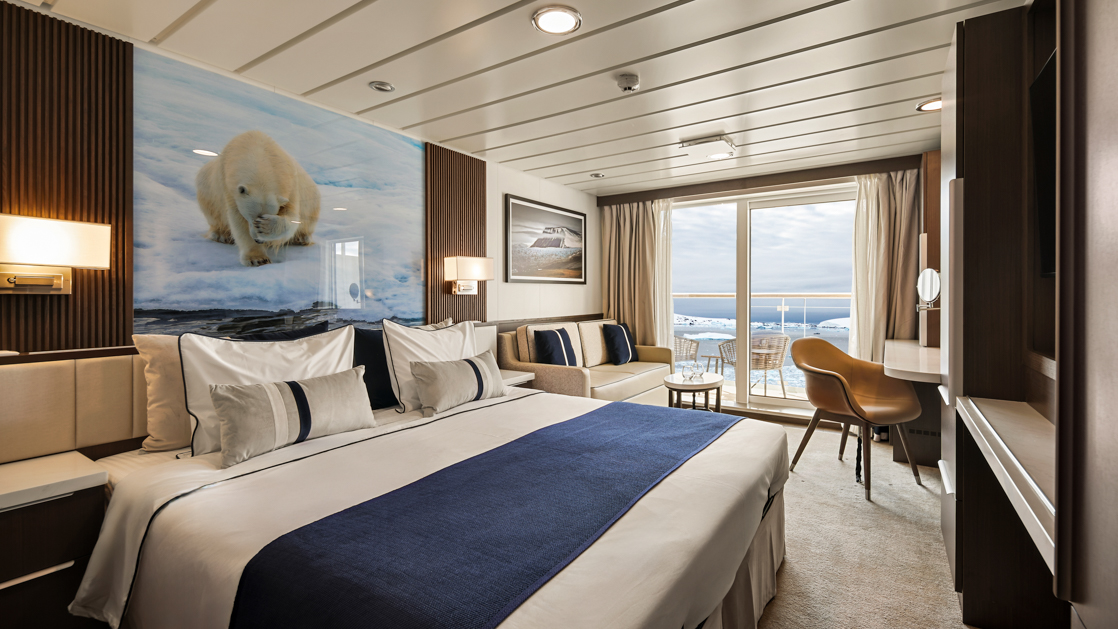 Category B Balcony Stateroom on Douglas Mawson ship with king bed in white linens, dark wood walls, balcony & wildlife photo.