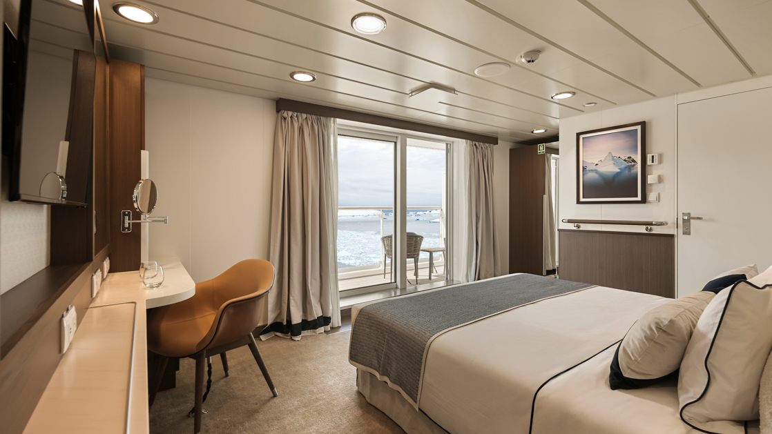 A balcony stateroom superior aboard the Douglas Mawson expedition ship featuring a bed, a small sofa, a desk with a chair