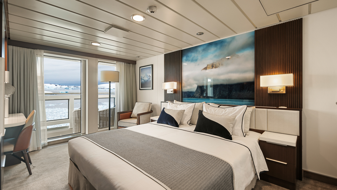 Captain's Suite bedroom on Douglas Mawson ship with king bed in white linens, dark wood walls, balcony & wildlife photo.