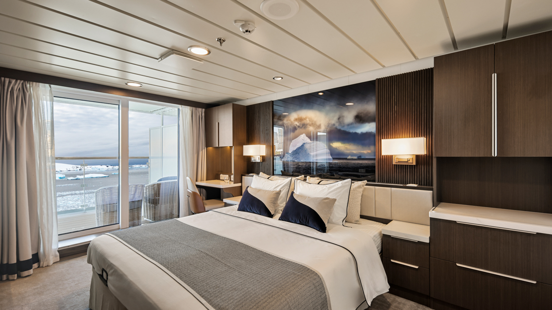 Junior Suite bedroom on Douglas Mawson ship with king bed in white linens, dark wood walls, balcony & wildlife photo.