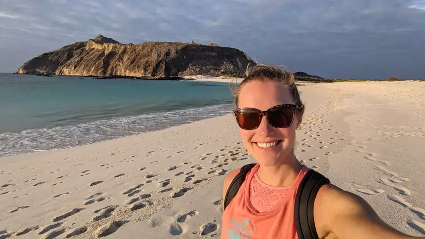Woman in coral-colored tank top & brown sunglasses stands on white-sand beach by teal sea in Galapagos.