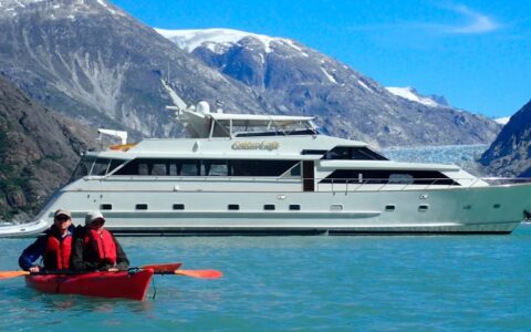 Two guests from the Golden Eagle yacht paddling in a double kayak in front of their small ship on a sunny day in Alaska.