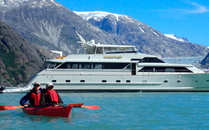 Two guests from the Golden Eagle yacht paddling in a double kayak in front of their small ship on a sunny day in Alaska.