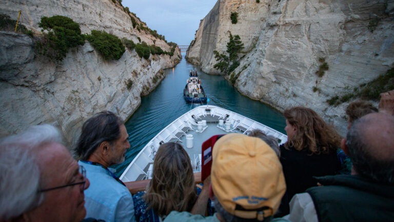 Travelers watch from the upper deck of a white small ship as a tug boat pulls them through the Corinth Canal's beige walls.
