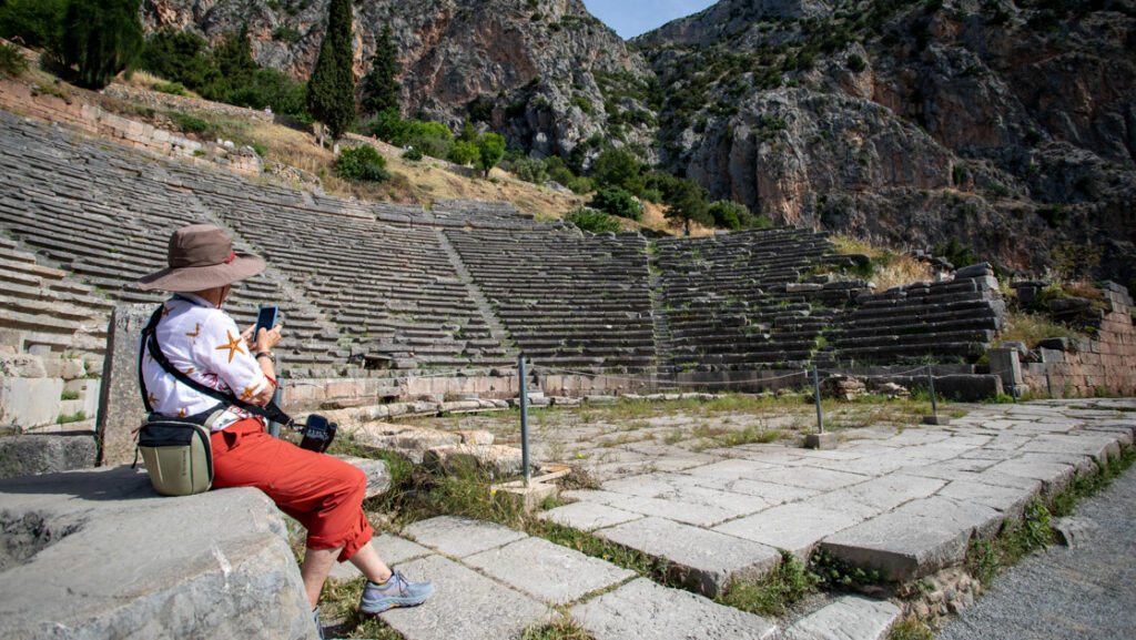 Greece traveler in red & white clothes sits on stone bench & photographs empty ancient amphitheater nestled among rocky cliffs.