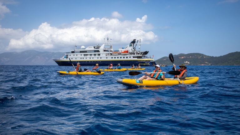 Travelers paddle inflatable yellow tandem kayaks near a National Geographic small ship in choppy blue sea on a sunny day.