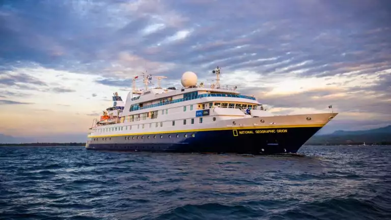 National Geographic Orion small ship with dark blue hull & white upper decks sits in calm water under a cloudy purple sky.