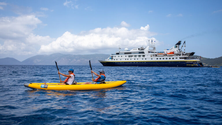 A pair of tandem kayakers in an inflatable yellow boat paddle calm blue sea beside the National Geographic Orion small ship.