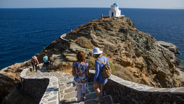 Greece travelers traverse a stone walkway along the spine of a cliff, towards a blue-domed, whitewashed church by the sea.