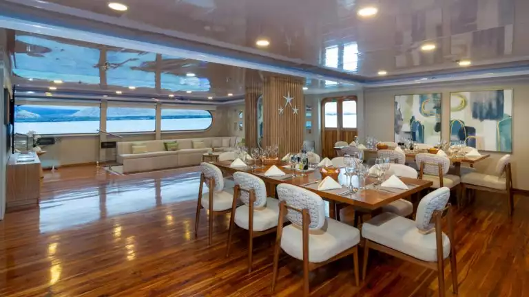 Dining area of Petrel catamaran in Galapagos with wood floor, 2 8-guest tables, dark wood chairs & marble food servicebar.