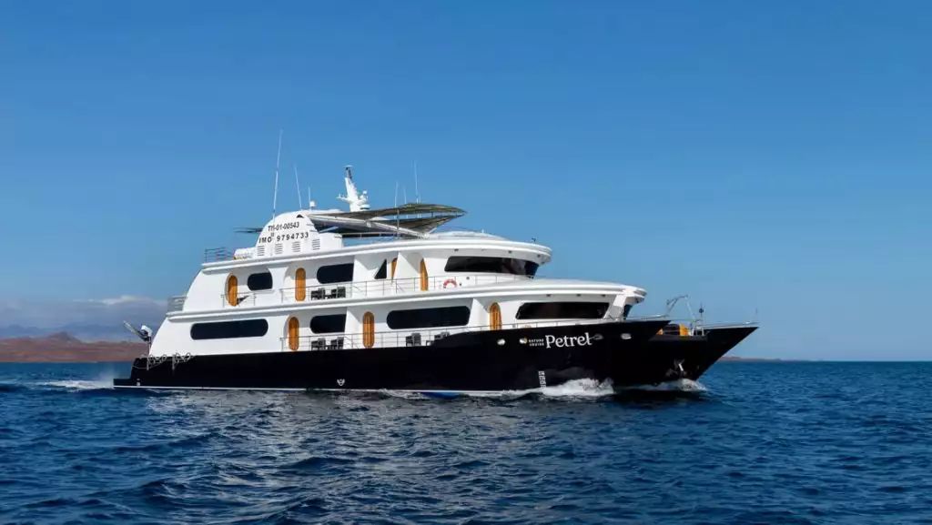 Petrel Galapagos catamaran with dark blue dual hull & 3 white upper decks cruises through calm water on a sunny day.