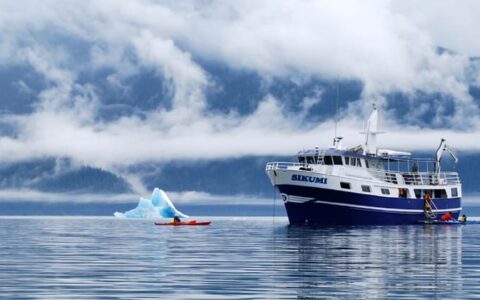 sikumi small ship anchored while an adventure traveler kayaks in front of an iceberg on a misty day in alaska