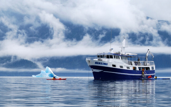 sikumi small ship anchored while an adventure traveler kayaks in front of an iceberg on a misty day in alaska