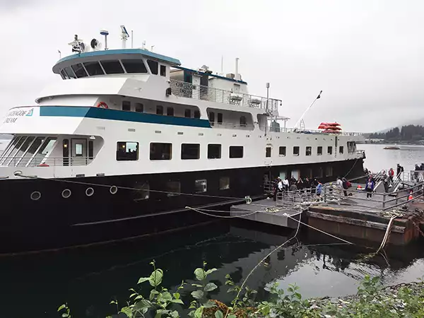A small ship with a black hull seen tied up next to a dock on a cloudy day with passengers loading on the ship.
