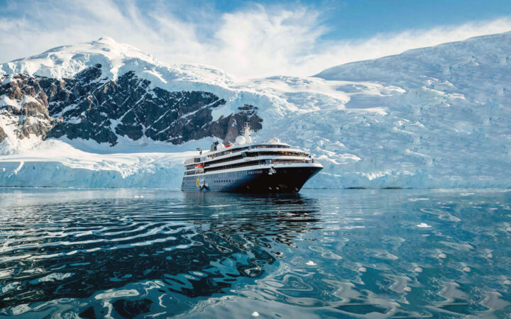 World Voyager cruise ship with blue hull & white upper decks sits in glassy Antarctic waters beside snowy peaks in the sun.
