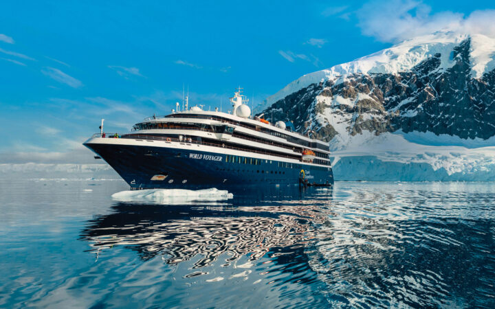 Small expedition ship World Voyager with blue hull & white upper decks sits in calm sea beside snowcapped peaks in Antarctica.