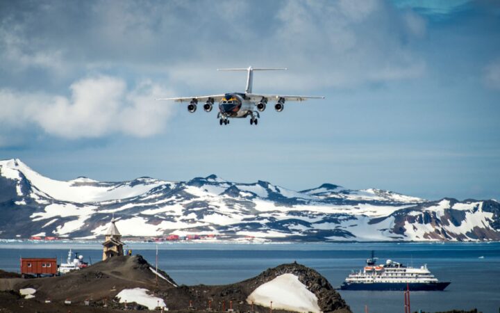 A plane comes in to land at Frei Station in Antarctica with blue skies and a ship in the ocean.