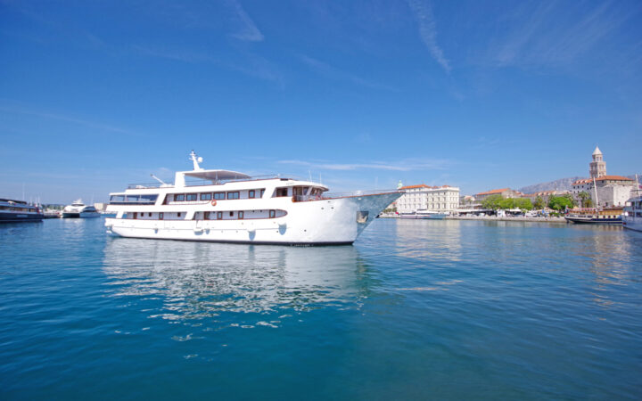 Aurora yacht with 4 guest decks & white exterior sits in calm turquoise water just offshore under sunny skies in Croatia.