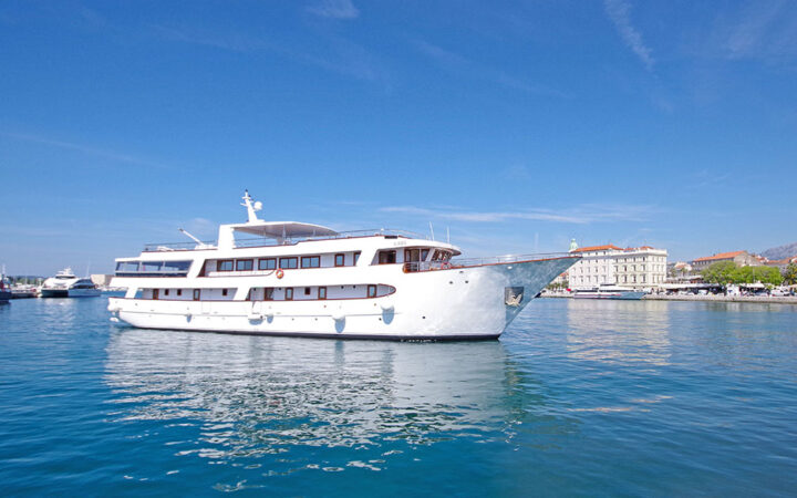 Aurora yacht with 4 guest decks & white exterior sits in calm turquoise water just offshore under sunny skies in Croatia.