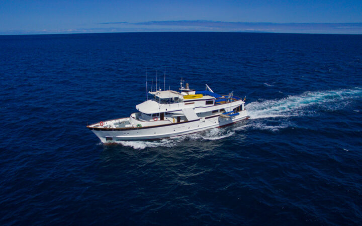 Aerial view of Beluga yacht with 3 passenger decks & white exterior cruising on a sunny day under blue sky in the Galapagos.