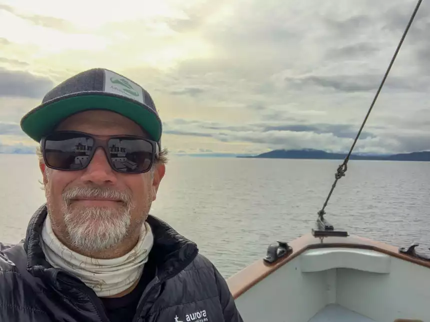 Man in gray hat, brown sunglasses & black puffy coat stands on deck near bow of Catalyst wooden ship on a cloudy day in Alaska.