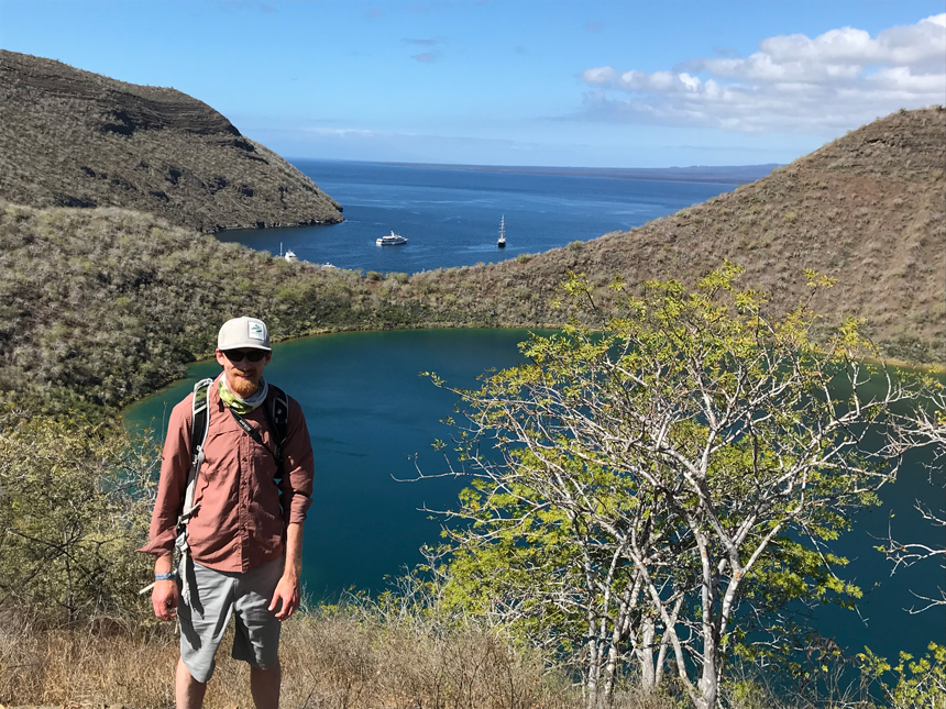 Man in red shirt, beige shorts & cap stands overlooking a small blue lake with sea beyond on a sunny day in Galapagos.
