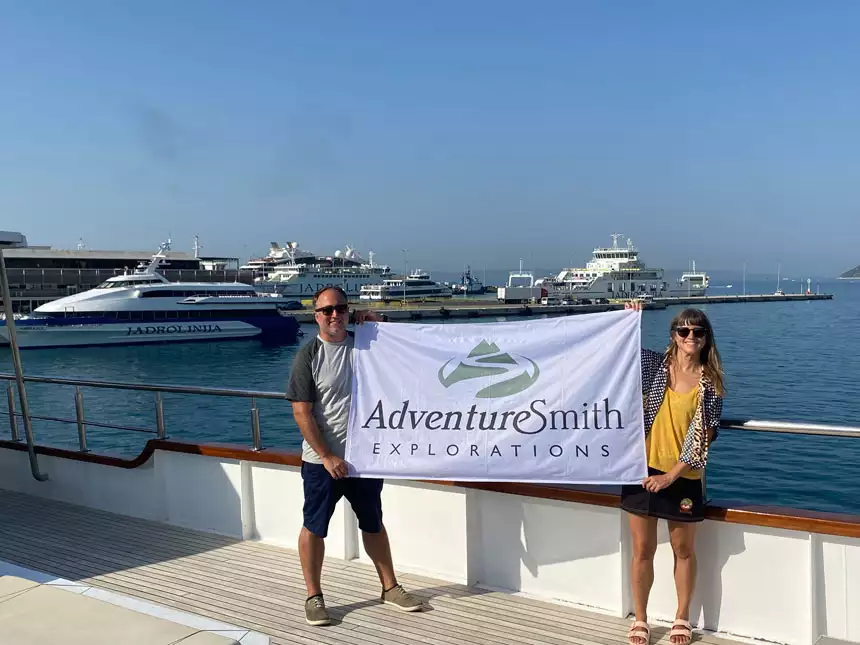 Man & woman hold a white & green AdventureSmith Explorations flag on open-air deck of M/S Aurora in Croatia.