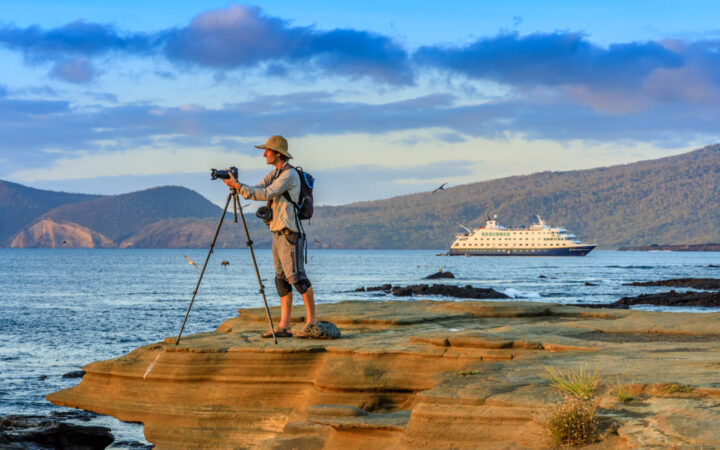 A photographer at sunset, on the Rocky Shoreline, with the ship National Geographic Endeavor II in the background, Santiago Island, Galapagos Islands, Galapagos National Park, Ecuador.
