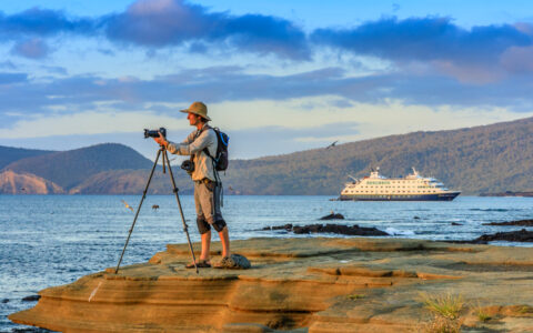 A photographer at sunset, on the Rocky Shoreline, with the ship National Geographic Endeavor II in the background, Santiago Island, Galapagos Islands, Galapagos National Park, Ecuador.
