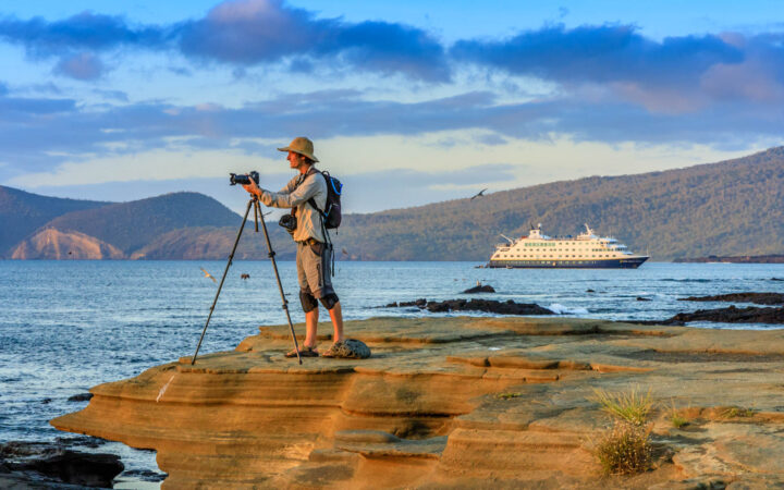A photographer at sunset, on the Rocky Shoreline, with the ship National Geographic Endeavor II in the background, Santiago Island, Galapagos Islands, Galapagos National Park, Ecuador.