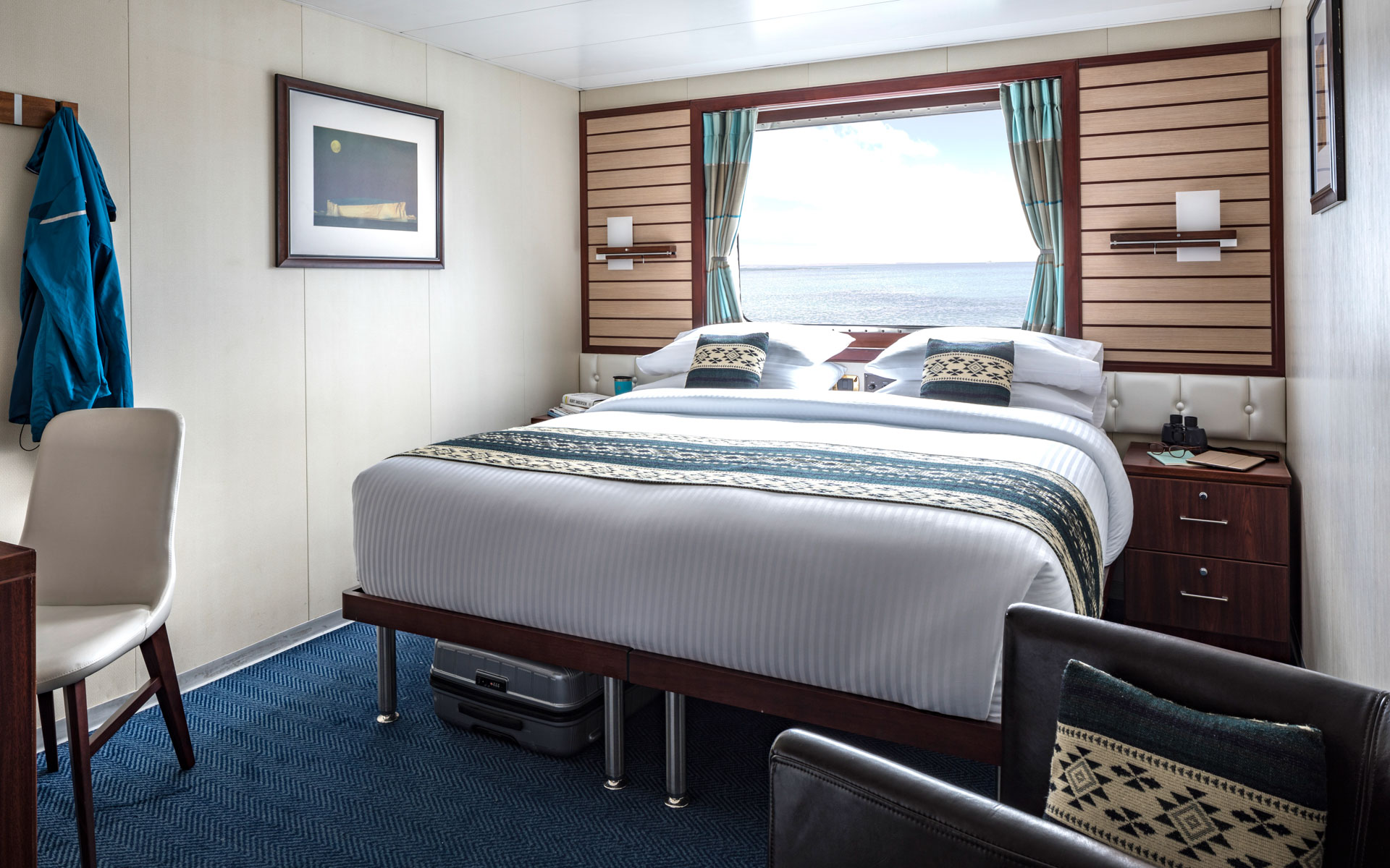 Category 2, 3 & 4 cabin interior with large bed, armchair, bedside tables, desk, chair and window aboard National Geographic Endeavour II expedition ship in the Galapagos Islands