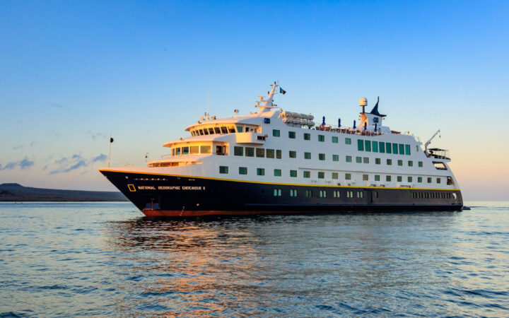 National Geographic Endeavour II ship with dark blue hull & white upper decks cruises through calm sea at sunset in Galapagos.