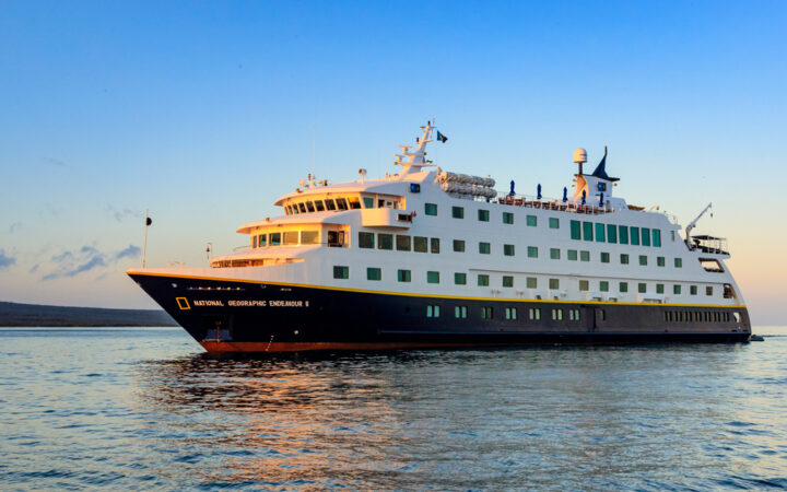 National Geographic Endeavour II ship with dark blue hull & white upper decks cruises through calm sea at sunset in Galapagos.