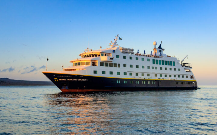 National Geographic Endeavour II ship with dark blue hull & white upper decks cruises through calm sea at sunset in Galapagos.