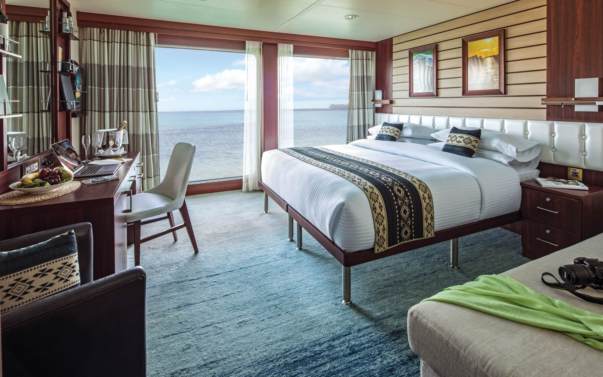 Interior of Suite B with large bed, couch, armchair, desk, chair and floor-to-ceiling windows aboard National Geographic Endeavour II expedition ship in the Galapagos Islands