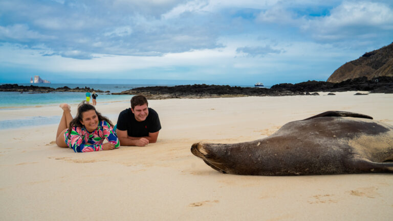 Ocean Spray Galapagos cruise guests lay on white-sand beach near a sea lion lounging on its back on a sunny day.