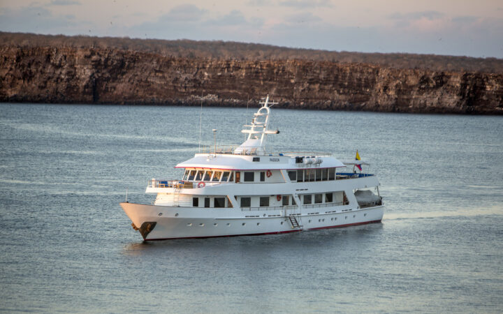 Passion luxury yacht with 4 guest decks, white exteriors & view windows cruising past brown island at sunset in Galapagos.