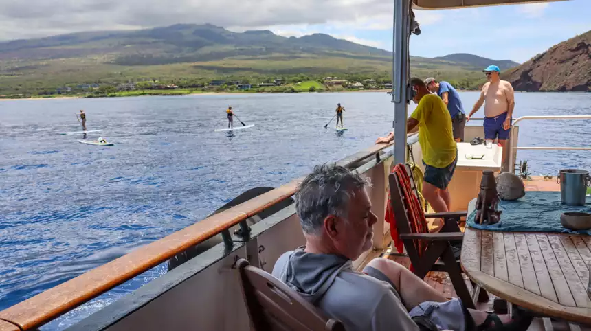 Guests aboard a small cruise ship on the deck looking out at paddle boarders 