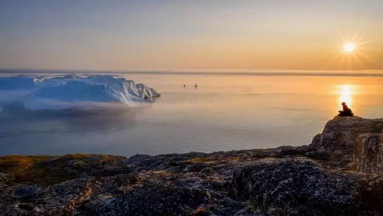 South Greenland Exploration cruise traveler sits alone on top of rocky cliff overlooking calm sea & snoy icebergs at sunset.