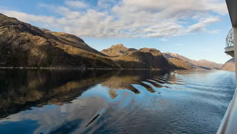 View from Nat Geo Endurance ship as she passes through Kuannersooq Fjord in Greenland with glassy water & rolling rocky hills.