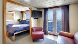 Safari Endeavour Alaska small ship Commodore suite with a large open room with glass doors leading to a balcony, a tv and a large bed in the adjoining room