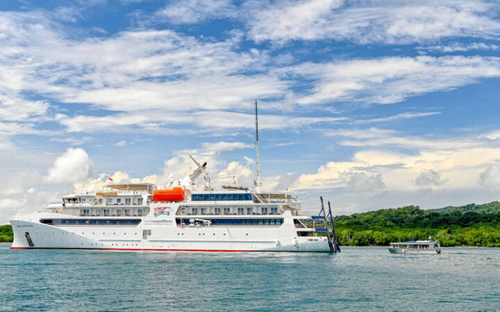 Exterior port side of Coral Geographer Australian expedition ship with gray metal tender vessel behind under blue sky.