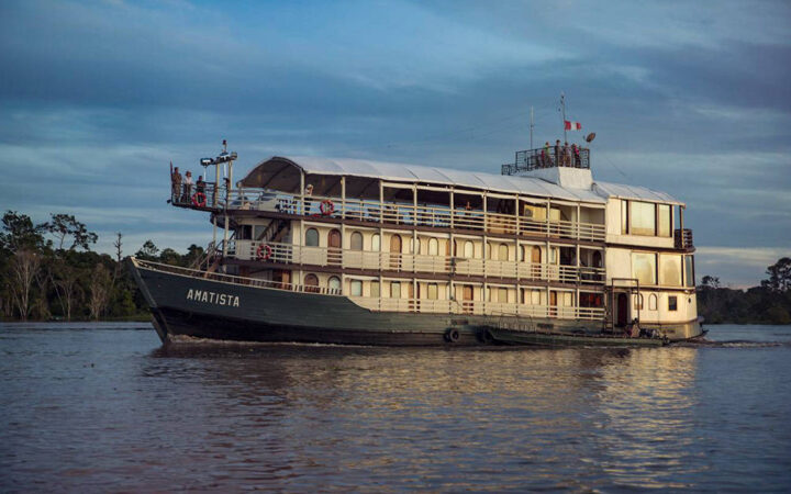 Wooden Amatista river boat with green hull & 3 white upper decks cruising at dusk in the Peruvian Amazon.