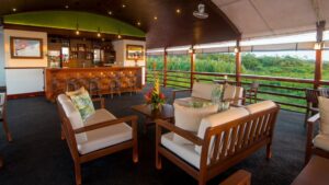 Upper deck, covered open-air bar on Amatista Amazon riverboat, with wood couches with white cushions, wood bar, fans & lighting.