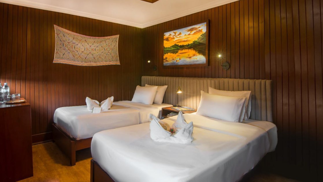 Ship cabin with 2 twin beds in white linens, dark wood paneled walls, plush gray headboard, desk & lamps on the Amatista.